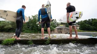 Surfer stehen an einem Wehr der Lahn. Eine künstliche Welle soll ihnen und anderen ermöglichen, den Wassersport fernab des Meeres auszuüben.
