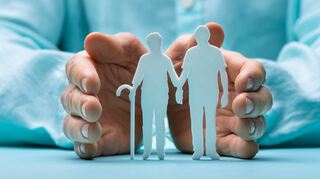 Hand Protecting Senior Couple Cutout Figures On Desk