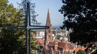 Ein stilisiertes Kreuz steht auf dem Freiburger Schlossberg während im Hintergrund das Münster zu sehen ist.