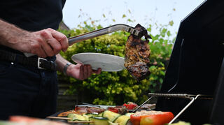 Eine Mann wendet mit einer Zange Grillfleisch auf einem Gasgrill. Symbolbild