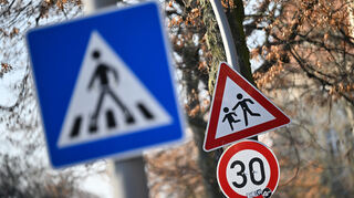 Ein Schild weist auf eine 30er-Zone und Kinder hin. Im Vordergrund ist ein Schild für einen Zebrastreifen zu sehen.