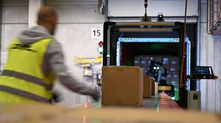 Ein DHL-Mitarbeiter nimmt Pakete in Emfpang - im Hintergrund ist ein Roboter zu sehen, der die Pakete aus einem Container nimmt und sie auf ein Förderband legt. (Archivbild)